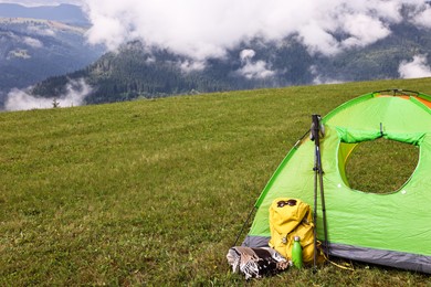 Tent, backpack, trekking poles and thermos on green grass in mountains, space for text