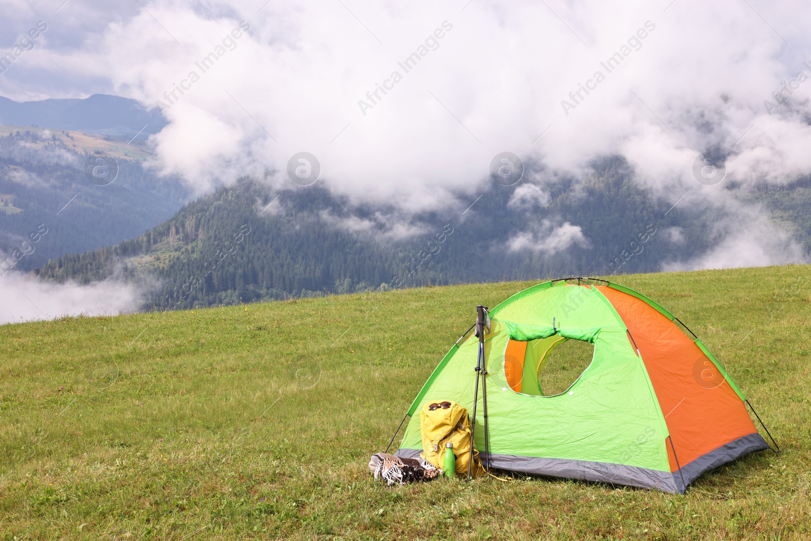 Photo of Tent, backpack, trekking poles and thermos on green grass in mountains, space for text