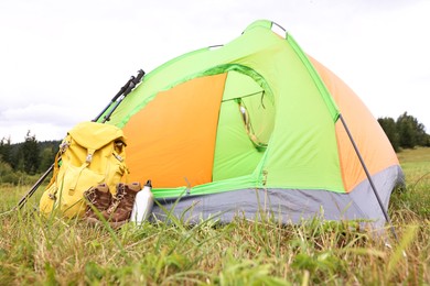 Tent, backpack, trekking poles and thermos on green grass in mountains