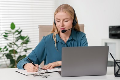 Photo of Interpreter in headset taking notes while working with laptop at table indoors