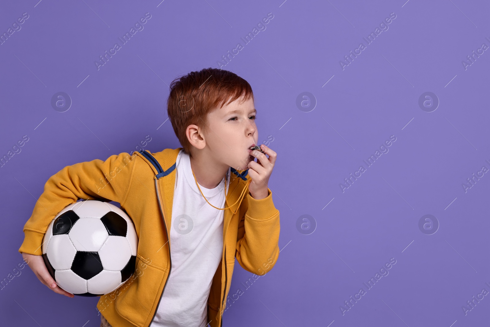 Photo of Little boy with soccer ball blowing whistle on purple background, space for text