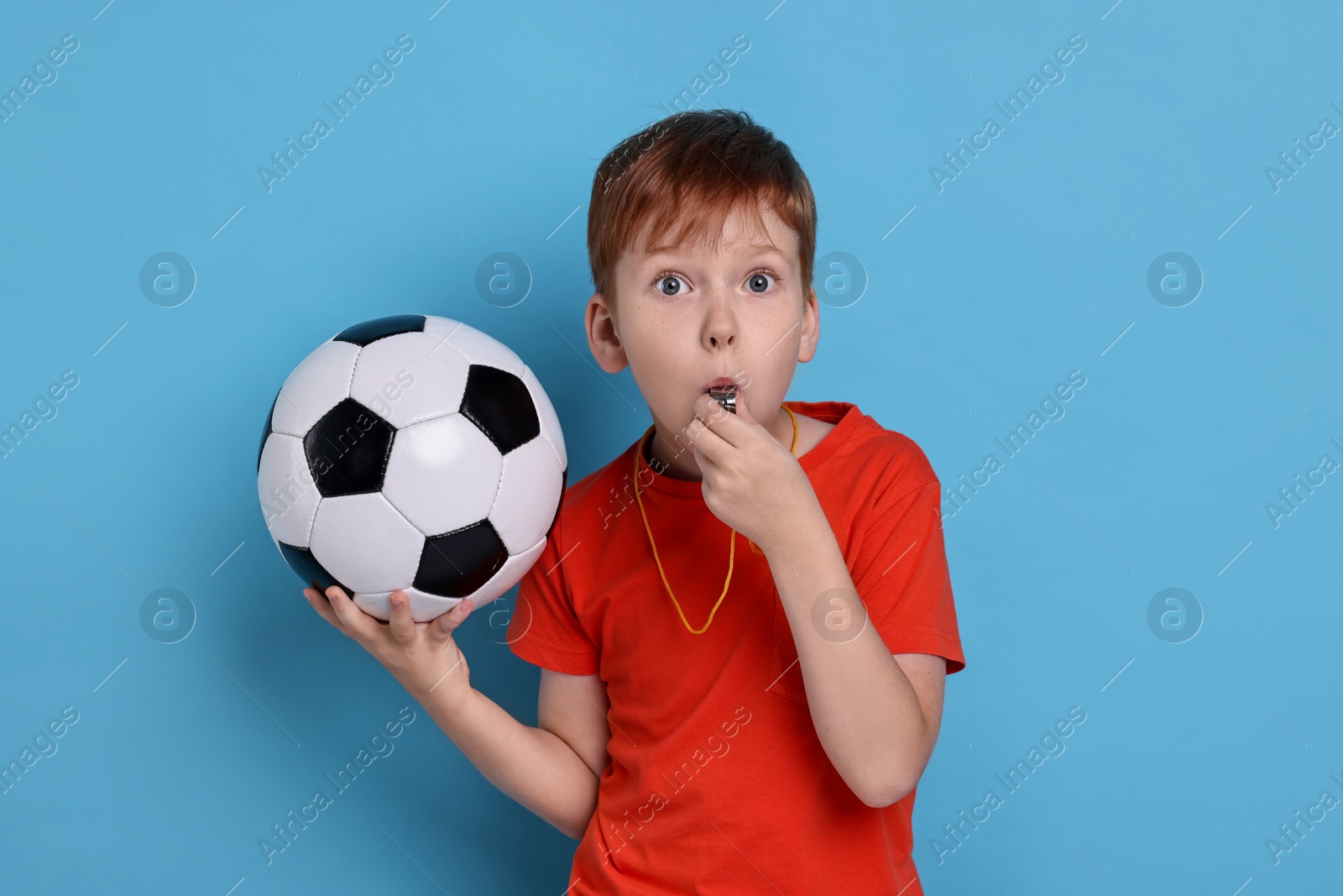 Photo of Little boy with soccer ball blowing whistle on light blue background