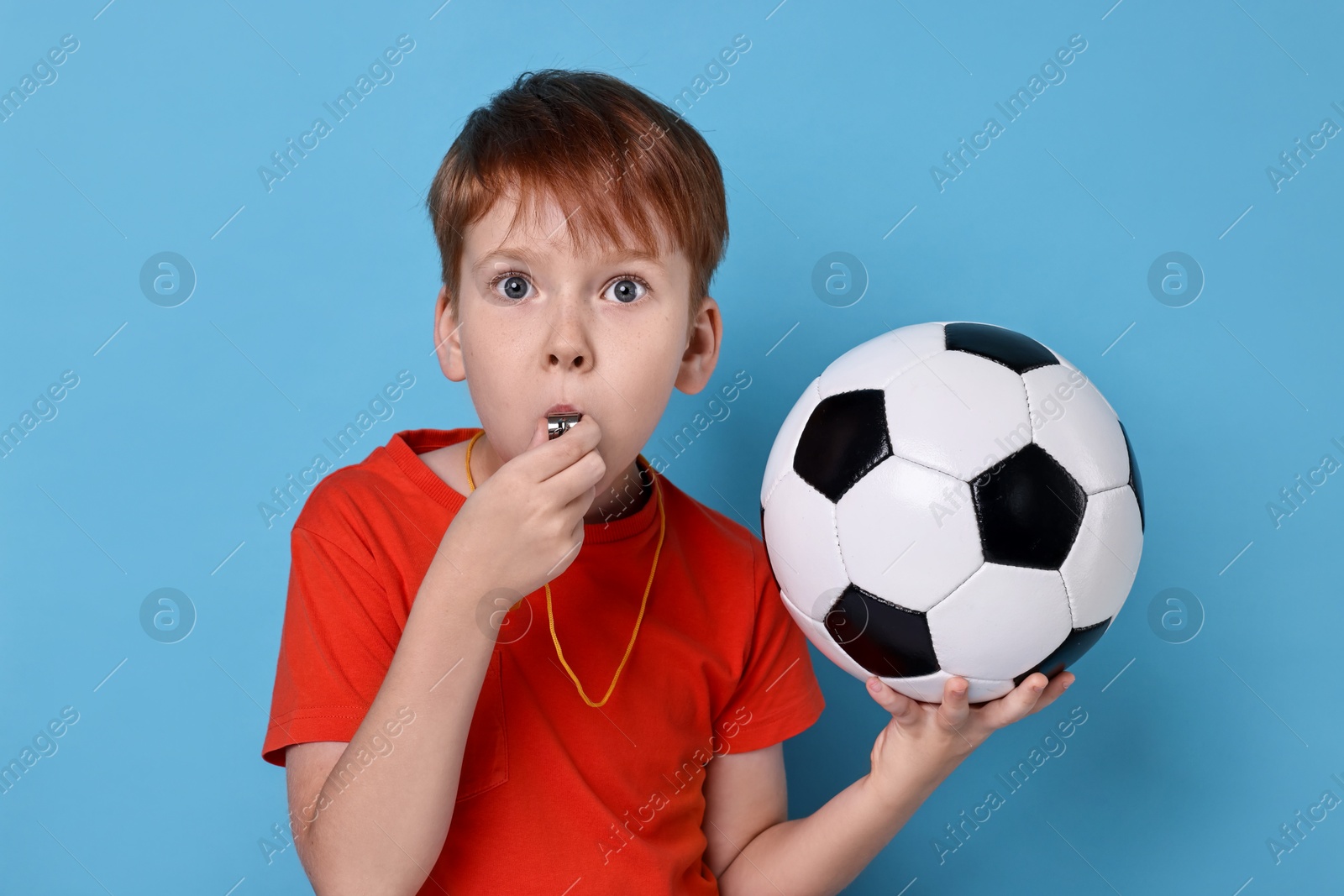 Photo of Little boy with soccer ball blowing whistle on light blue background