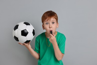 Little boy with soccer ball blowing whistle on grey background