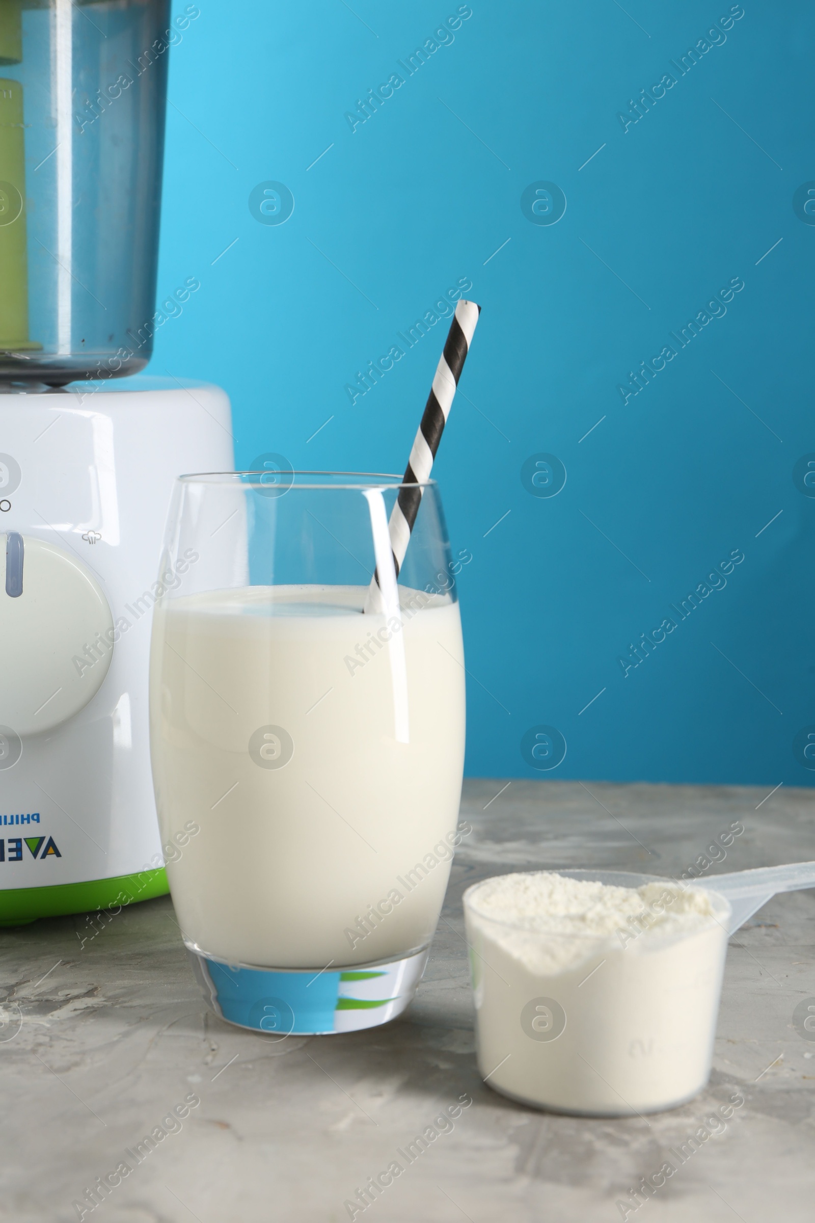 Photo of Delicious protein shake in glass, scoop with powder and blender on gray textured table against light blue background