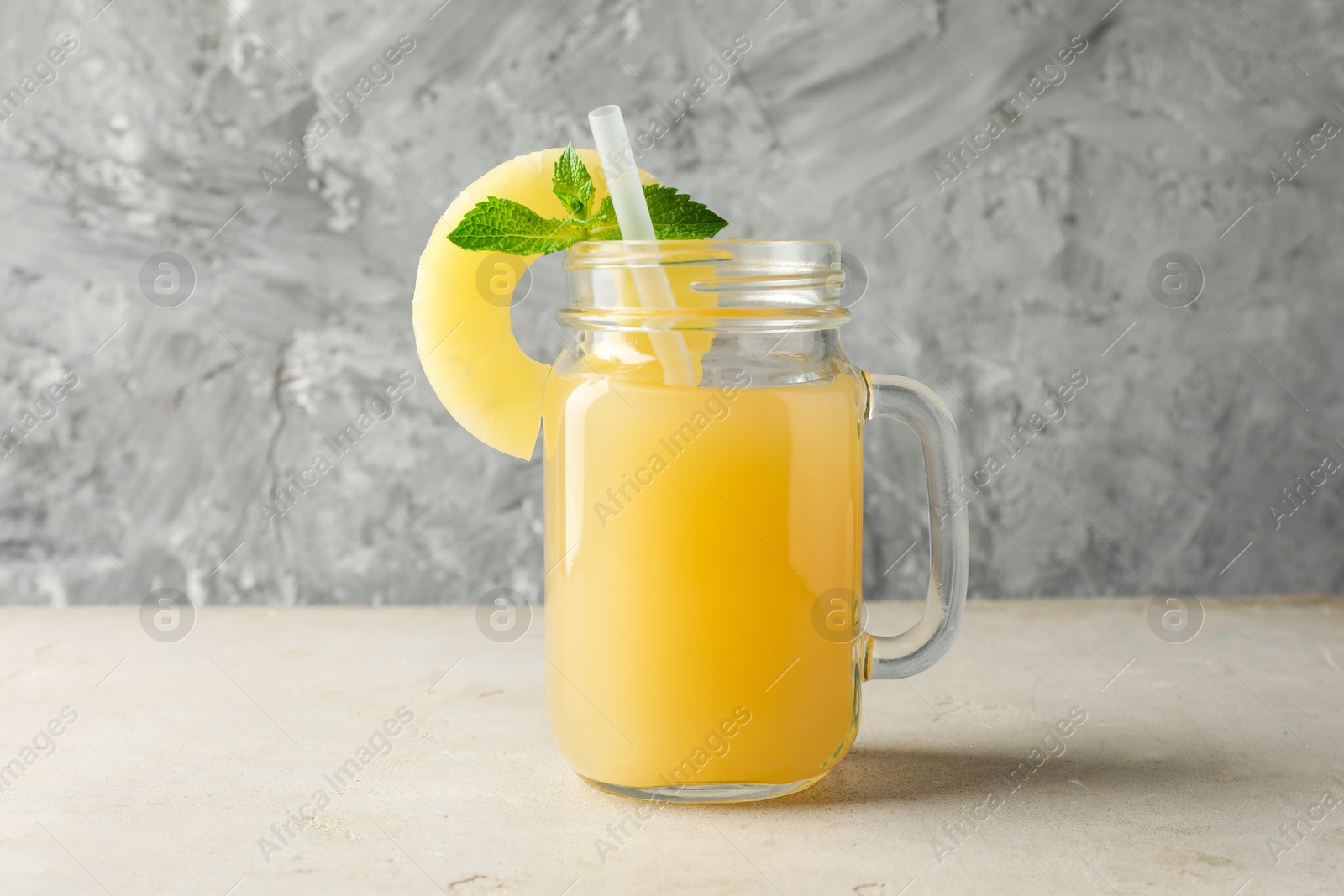 Photo of Tasty pineapple juice in mason jar, mint and slice of fresh fruit on grey textured table, closeup