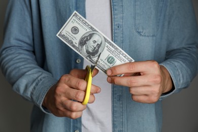 Photo of Budgeting. Man cutting dollar banknote with scissors on dark grey background, closeup