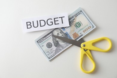 Photo of Dollar banknotes, scissors and paper with word Budget on white background, top view