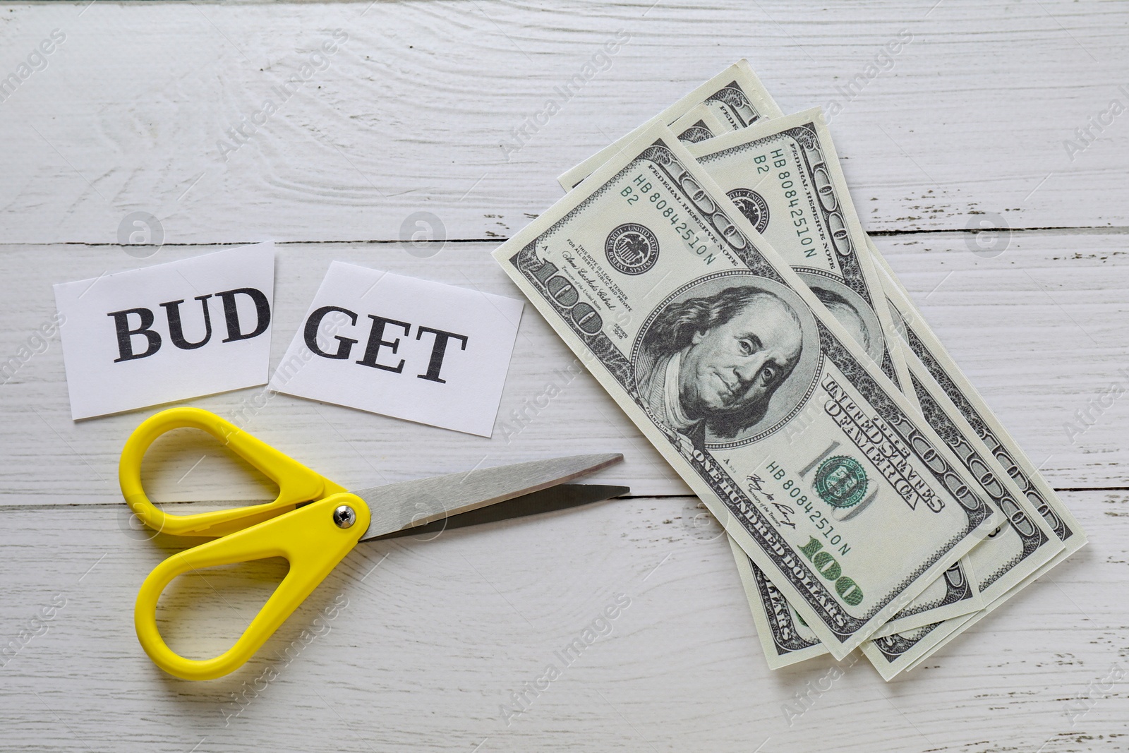 Photo of Cut paper with word Budget, dollar banknotes and scissors on white wooden table, top view