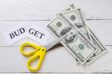 Photo of Cut paper with word Budget, dollar banknotes and scissors on white wooden table, top view