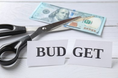 Photo of Cut paper with word Budget, dollar banknote and scissors on white wooden table, closeup