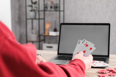 Photo of Online poker. Man with playing cards, chips and laptop at wooden table indoors, closeup