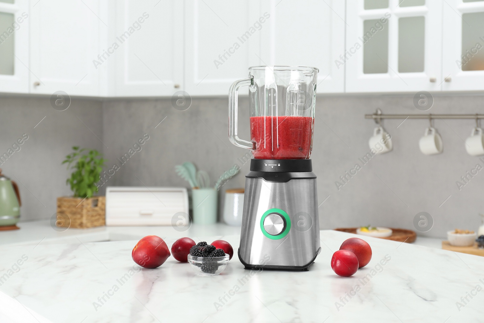 Photo of Blender with mixture of ingredients, fresh berries and fruits on white marble table indoors