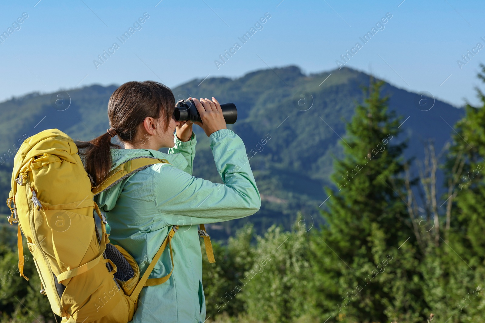 Photo of Young woman with backpack looking through
binoculars in beautiful mountains, space for text