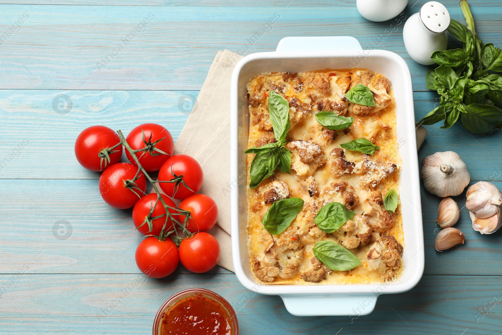 Photo of Tasty baked cauliflower in baking dish, basil, garlic, tomatoes and sauce on light blue wooden table, top view