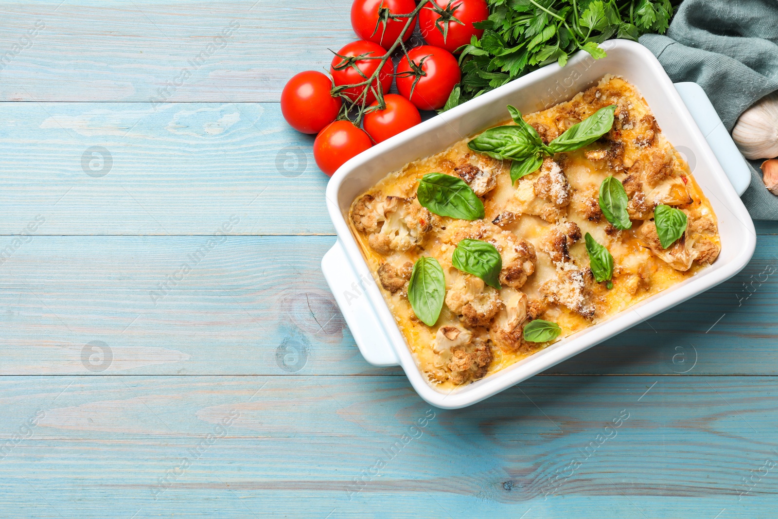 Photo of Tasty baked cauliflower in baking dish, basil, parsley, garlic and tomatoes on light blue wooden table, top view. Space for text