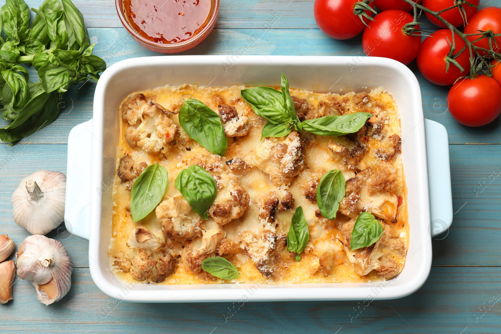 Photo of Tasty baked cauliflower in baking dish, basil, tomatoes, garlic and sauce on light blue wooden table, top view