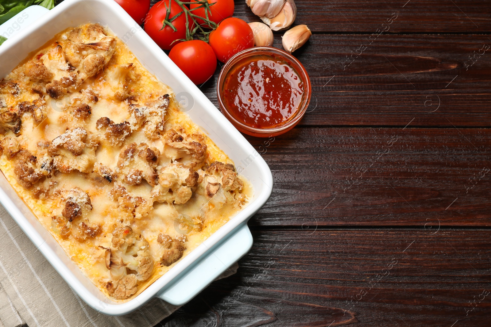 Photo of Tasty baked cauliflower in baking dish, tomatoes, sauce and garlic on wooden table, top view. Space for text