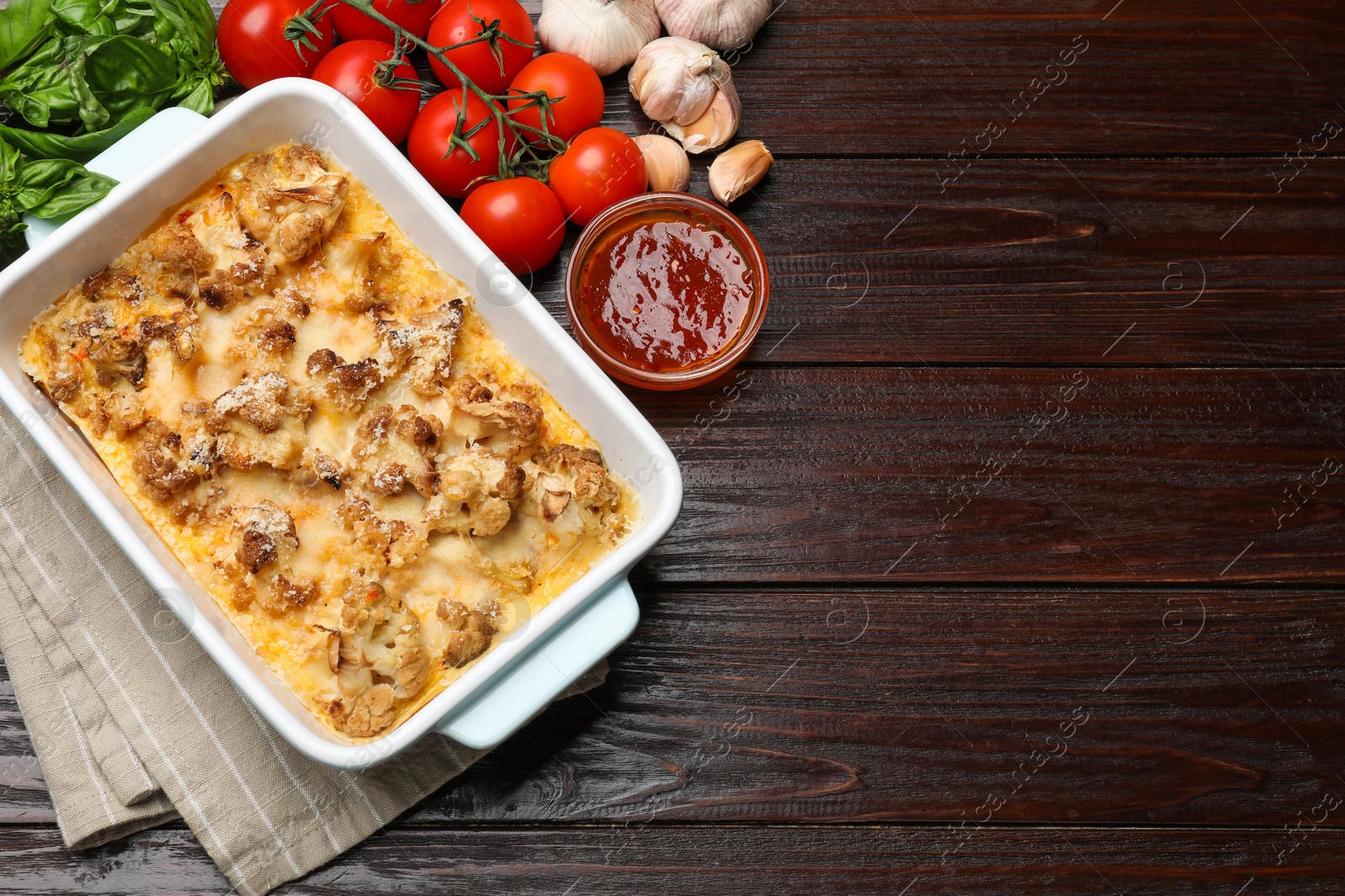 Photo of Tasty baked cauliflower in baking dish, basil, tomatoes, sauce and garlic on wooden table, top view. Space for text