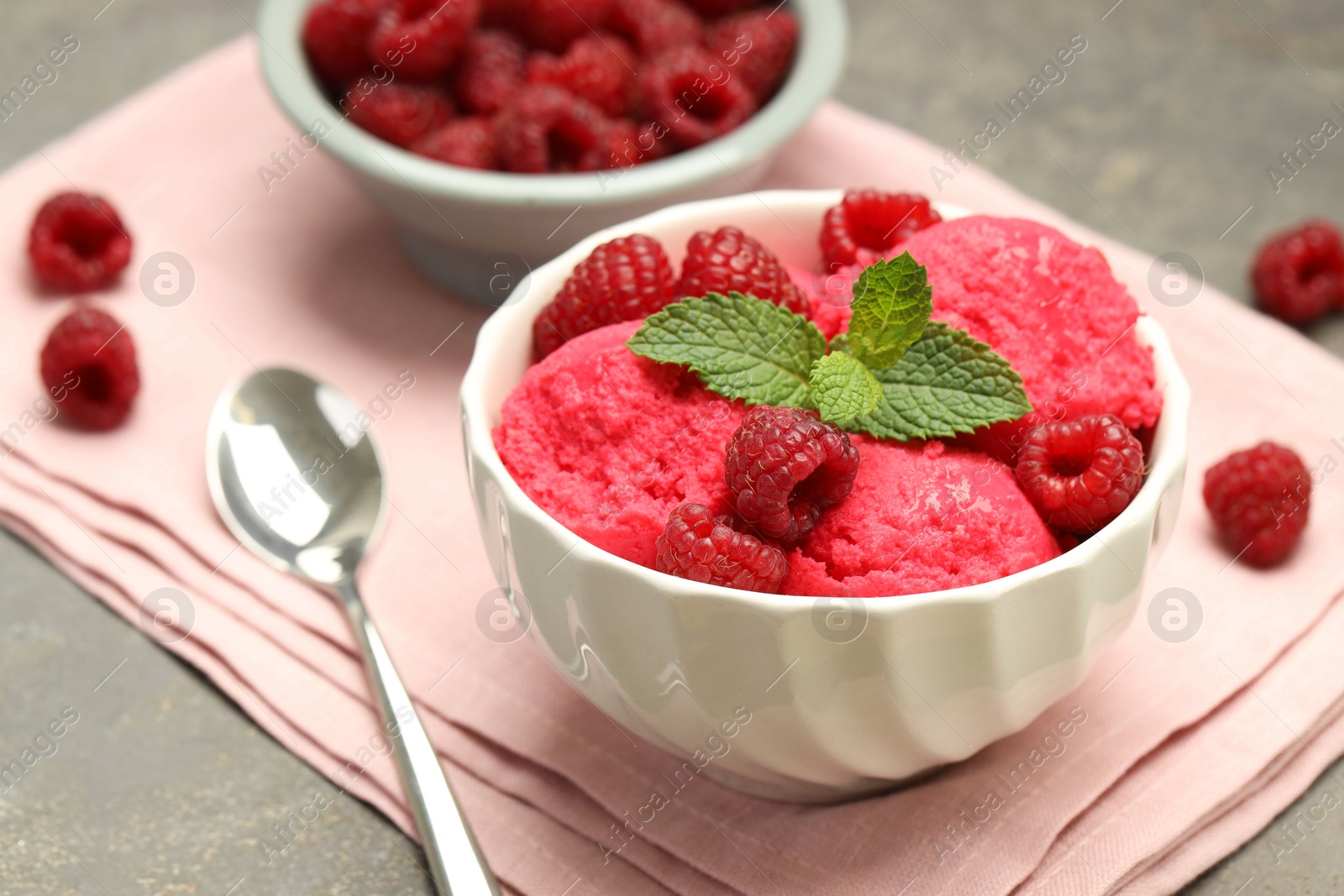 Photo of Delicious raspberry sorbet with fresh berries in bowl and spoon on gray textured table, closeup