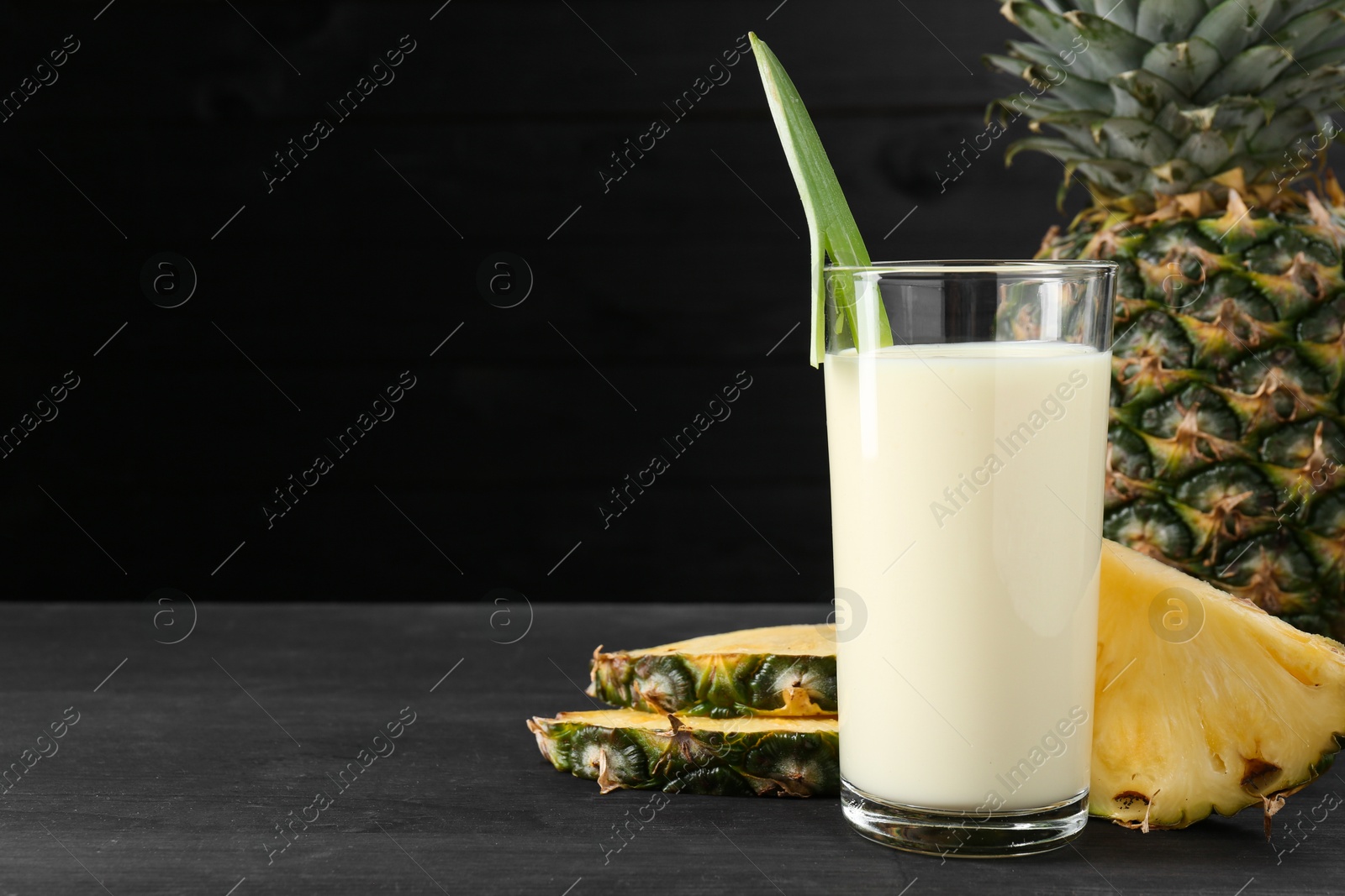 Photo of Tasty pineapple smoothie in glass and fruit on black wooden table, closeup. Space for text