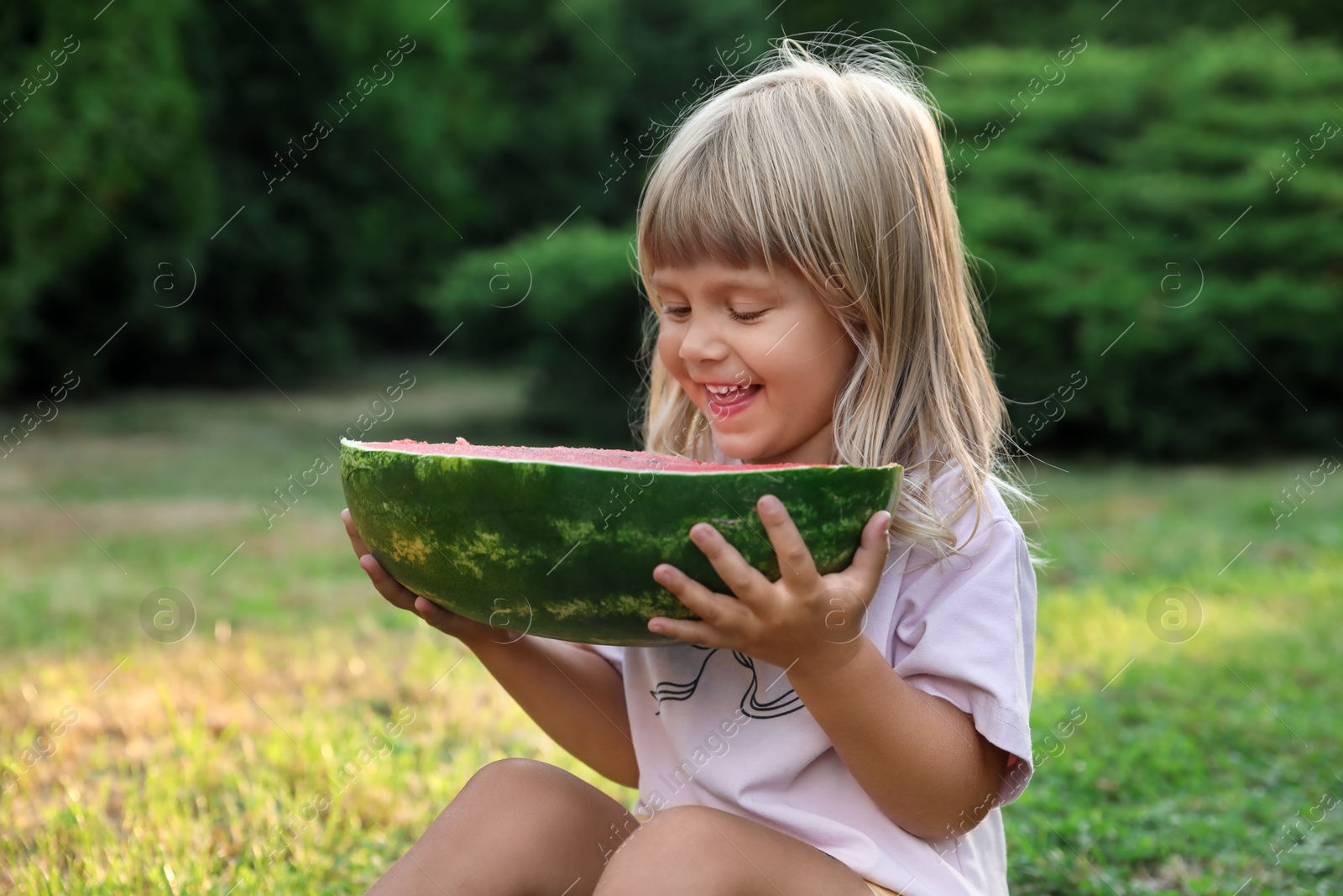 Photo of Cute little girl with half of juicy watermelon on green grass outdoors