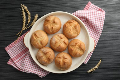 Photo of Baking dish with homemade tasty buns and spikelets on black wooden table, flat lay