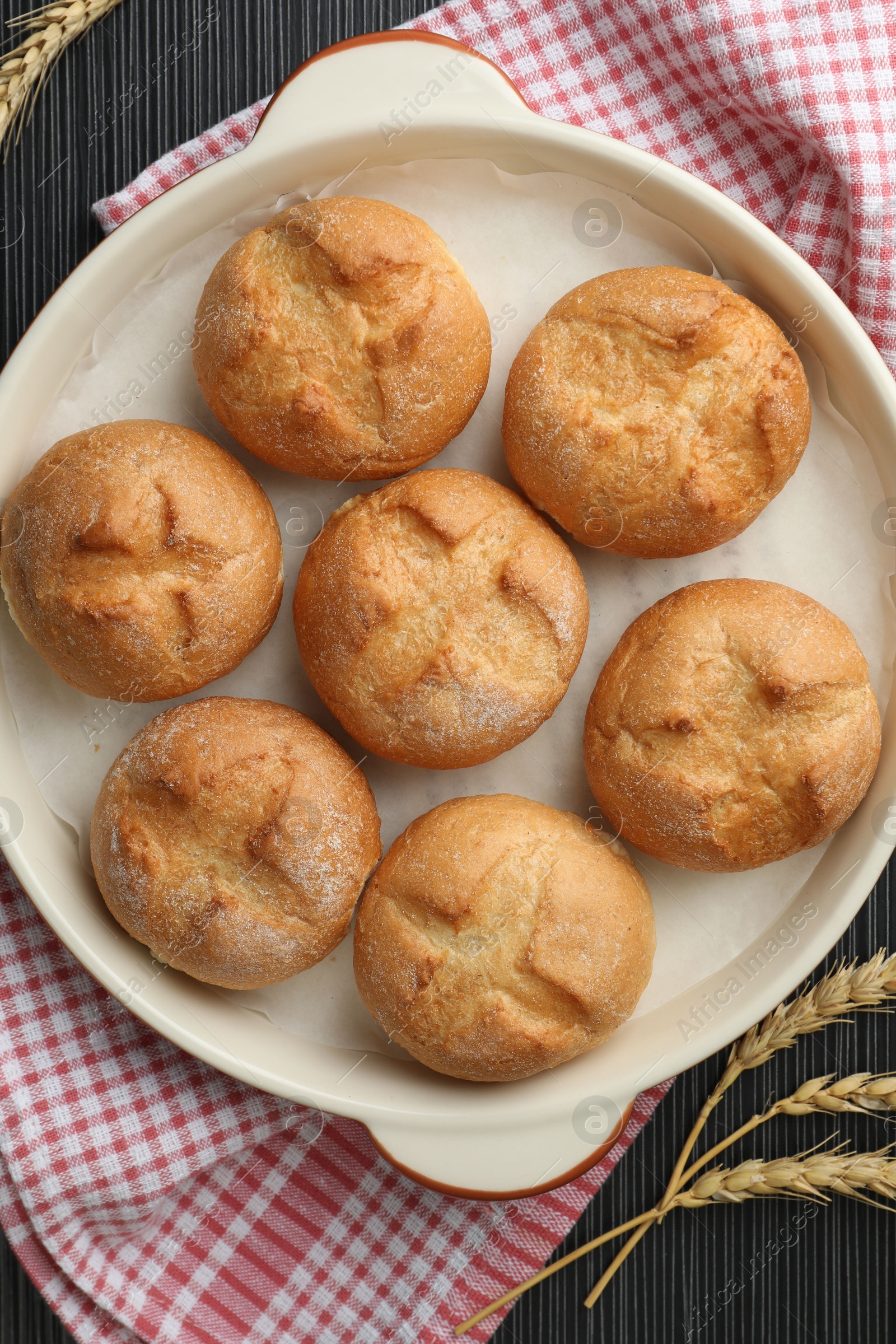 Photo of Baking dish with homemade tasty buns and spikelets on black wooden table, flat lay