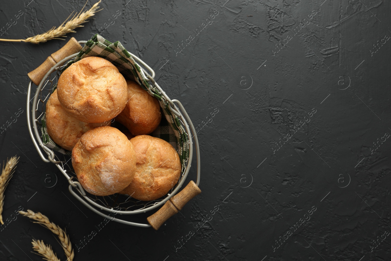 Photo of Metal basket with homemade tasty buns and spikelets on black textured table, flat lay. Space for text