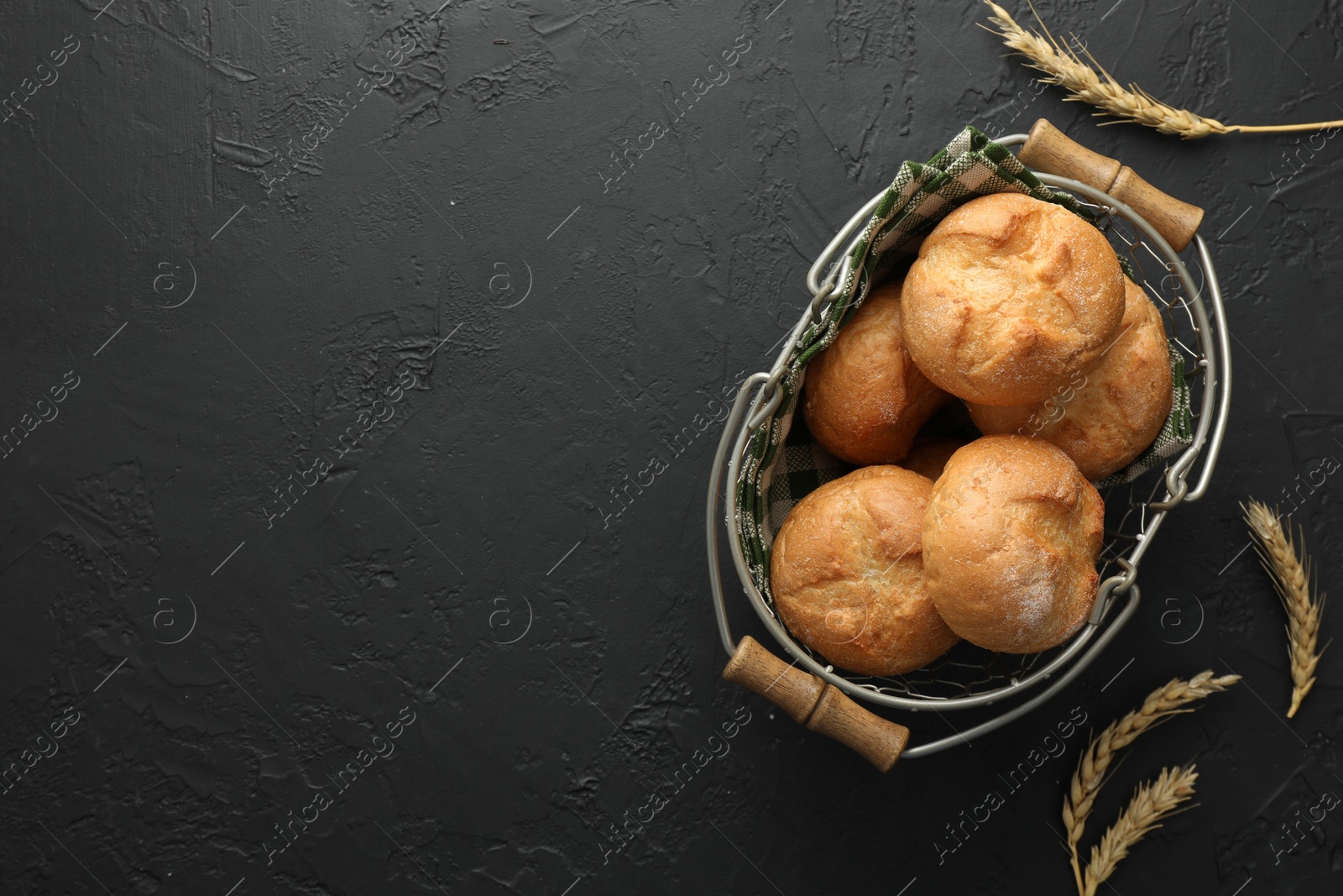 Photo of Metal basket with homemade tasty buns and spikelets on black textured table, flat lay. Space for text