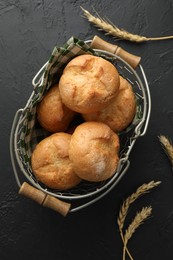 Photo of Metal basket with homemade tasty buns and spikelets on black textured table, flat lay