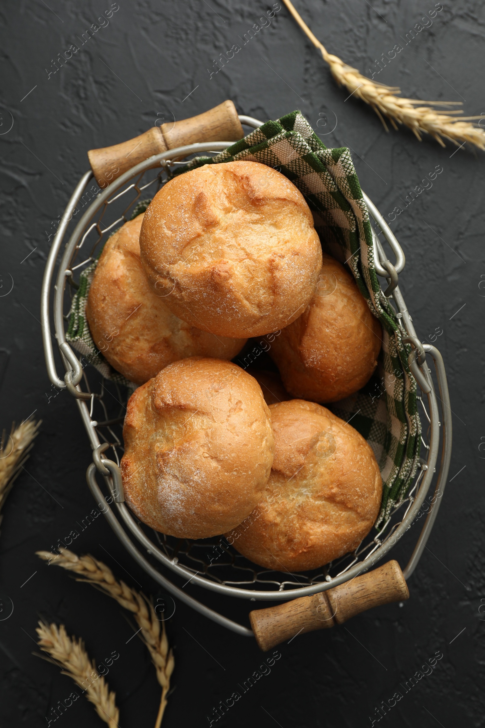 Photo of Metal basket with homemade tasty buns and spikelets on black textured table, flat lay