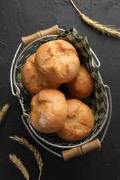 Photo of Metal basket with homemade tasty buns and spikelets on black textured table, flat lay