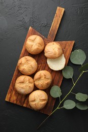 Homemade tasty buns and eucalyptus branch on black textured table, flat lay