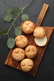 Photo of Homemade tasty buns and eucalyptus branch on black textured table, flat lay