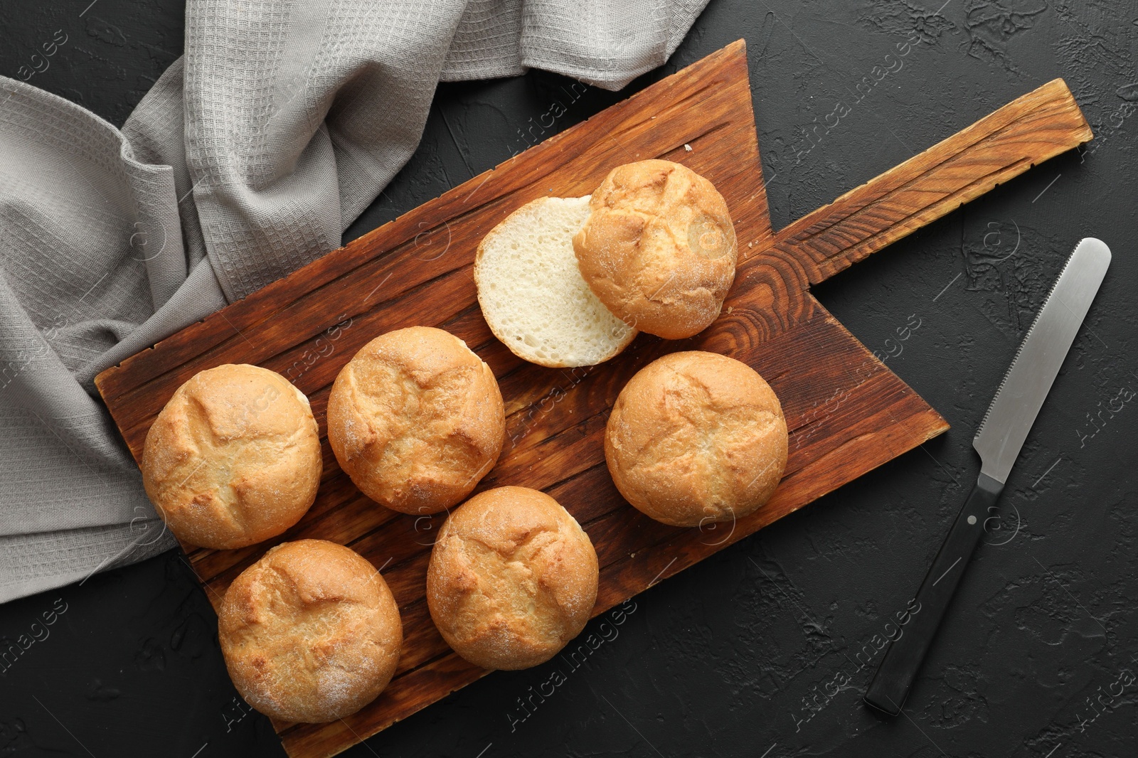 Photo of Board with homemade tasty buns and knife on black textured table, flat lay