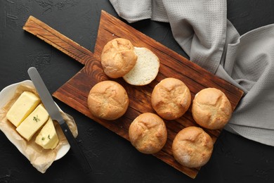 Board with homemade tasty buns, butter and knife on black textured table, flat lay