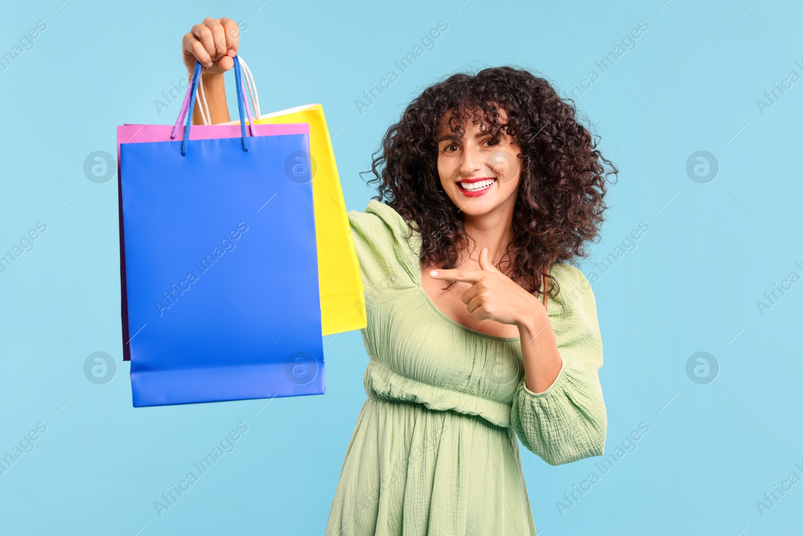 Photo of Smiling woman pointing at colorful shopping bags on light blue background