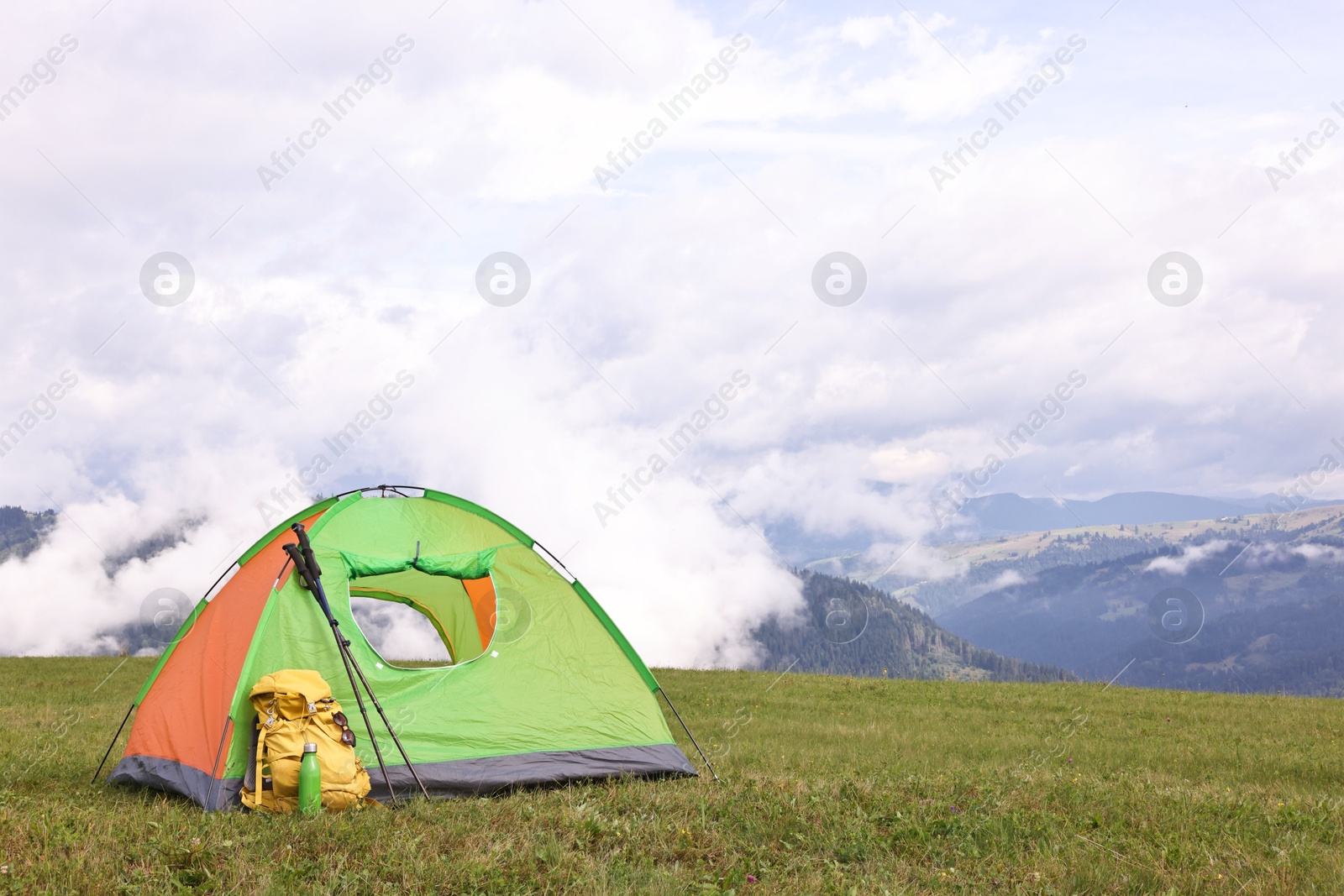 Photo of Tent, backpack, trekking poles and thermos on green grass in mountains, space for text