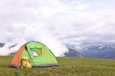 Tent, backpack, trekking poles and thermos on green grass in mountains, space for text