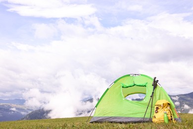 Tent, backpack, trekking poles and thermos on green grass in mountains, space for text