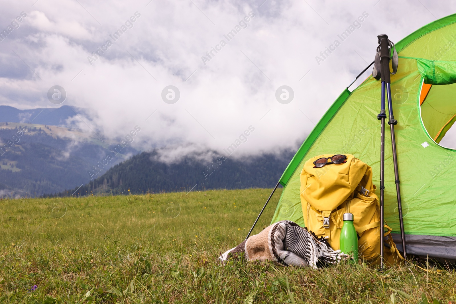 Photo of Tent, backpack, trekking poles and thermos on green grass in mountains, space for text