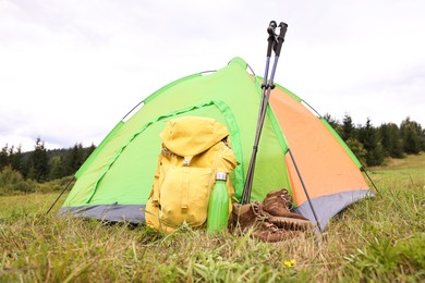 Tent, backpack, trekking poles and thermos on green grass in mountains