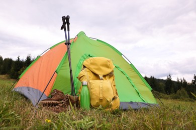 Tent, backpack, trekking poles and thermos on green grass in mountains