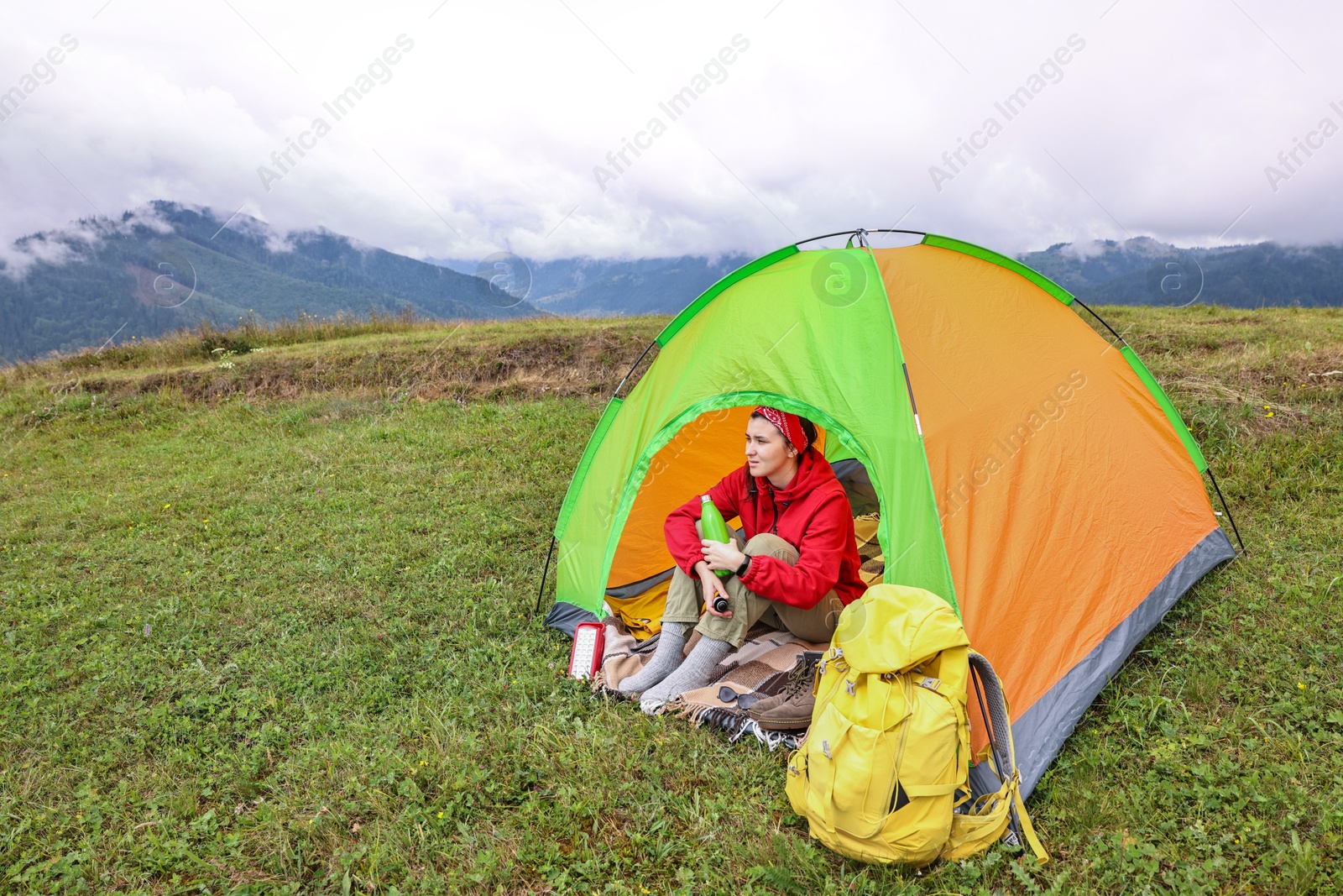 Photo of Young woman with thermo bottle sitting in camping tent on green grass outdoors, space for text