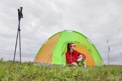 Young woman with thermo bottle sitting in camping tent on green grass outdoors