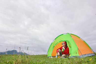 Photo of Young woman with thermo bottle sitting in camping tent on green grass outdoors, space for text