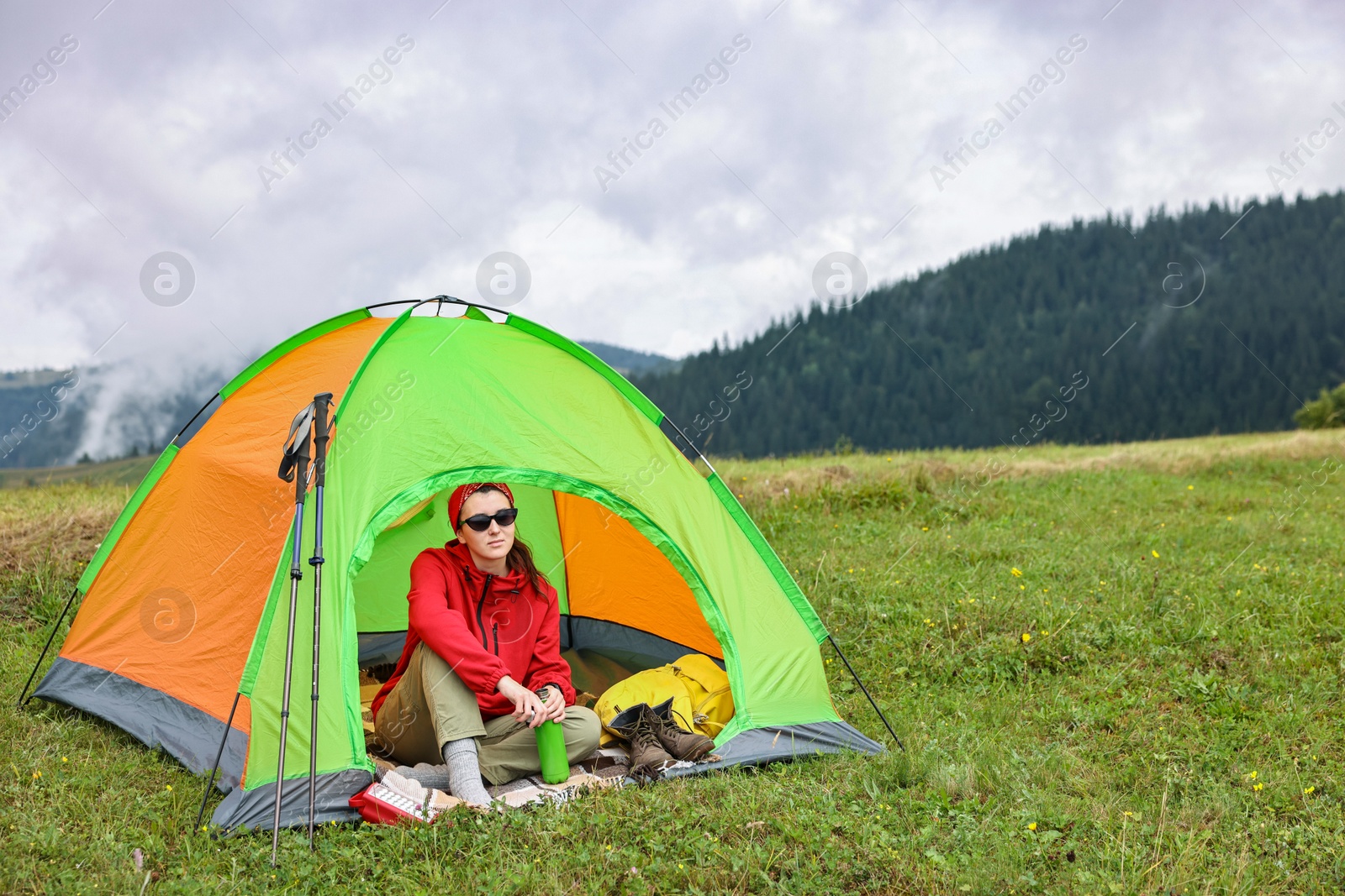 Photo of Young woman with thermo bottle sitting in camping tent on green grass outdoors, space for text