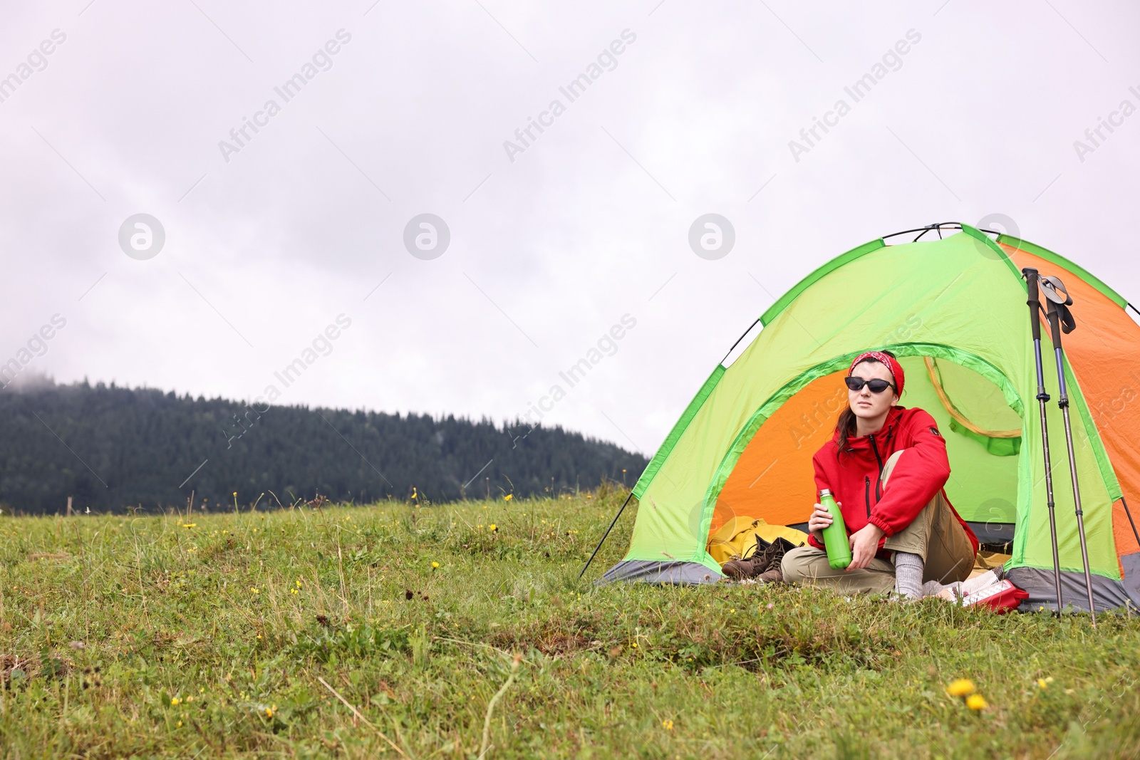 Photo of Young woman with thermo bottle sitting in camping tent on green grass outdoors, space for text