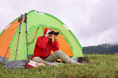 Young woman sitting in tent and looking through binoculars on green grass outdoors